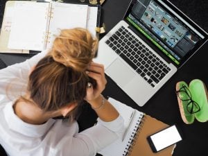 a person holding her head over a laptop and two notebooks, and a phone