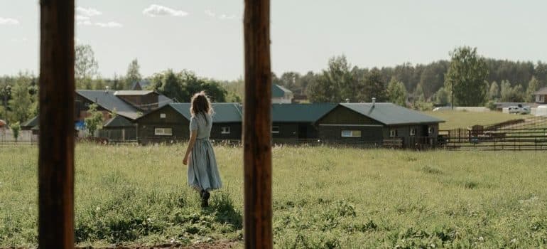 a woman in a dress standing on grass and looking at a farmhouse