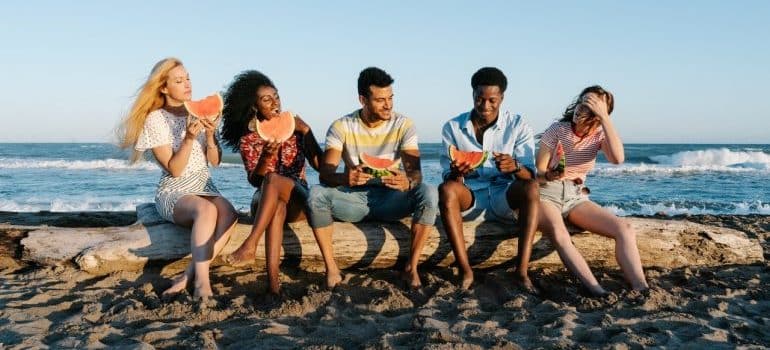 cheerful friends enjoying watermelon slices on sandy sea shore