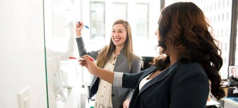 Two woman writing on a dry erase board