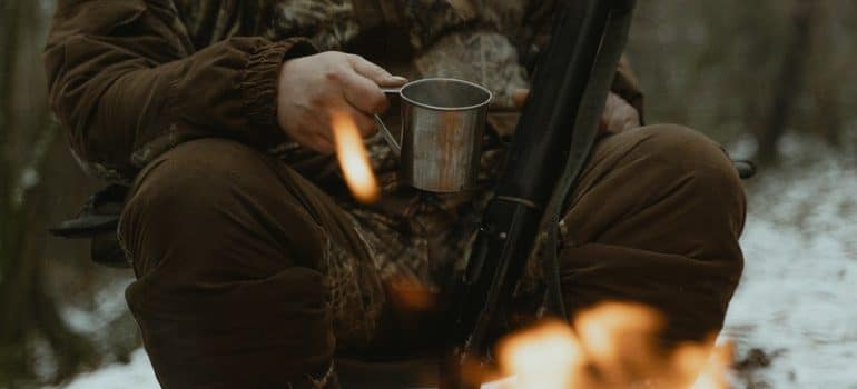 A faceless hunter siting by the fire with a metal mug and a shotgun between his legs.