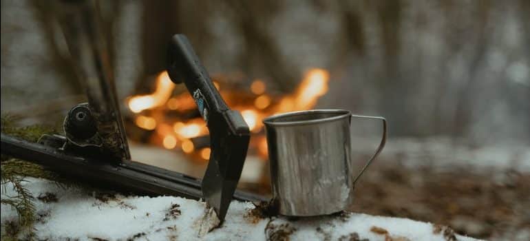 A hunting tool and a metal mug on a piece of wood in front of a fire in the snowy woods.