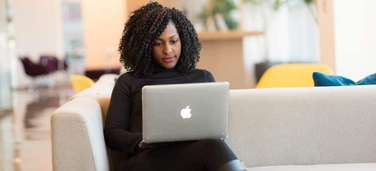 A women sitting on a white couch with a laptop on her lap thinking when is the best time to contact professionals when moving to Miami Beach.