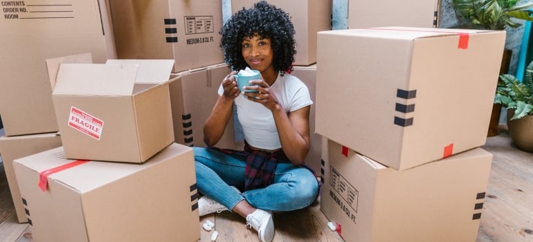 a woman sitting in between cardboard boxes while drinking a hot beverage