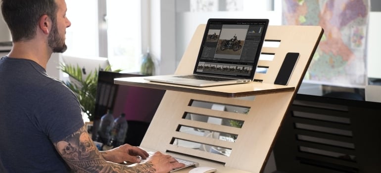 Man is standing and working on laptop placed at the adjustable office desk.