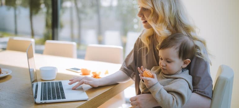 A woman is carrying a baby while browsing the internet on her laptop.