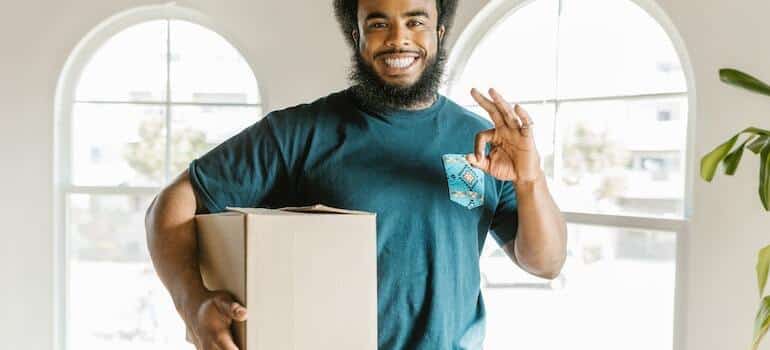 A smiling mover holding a moving box smiling knowing that hiring antique moving services is convenient