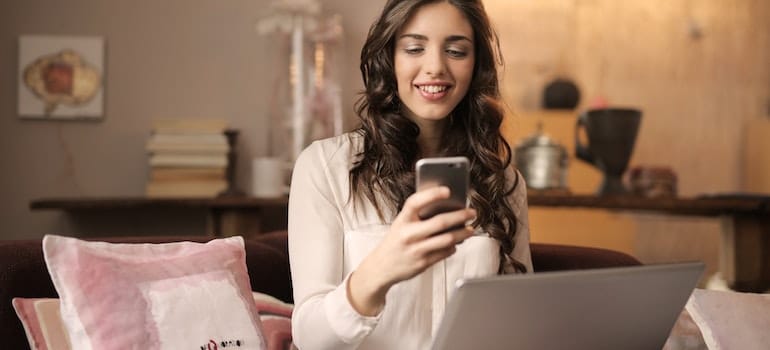 Woman Sitting on Sofa While Looking at Phone With Laptop on Lap - asking, you can easily get moving discounts in October. 