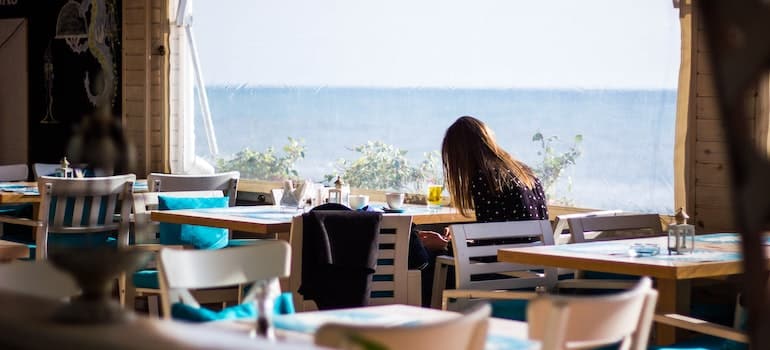 A woman sitting in a chair in a restaurant next to a window, while thinking moving to Pompano Beach around Thanksgiving;