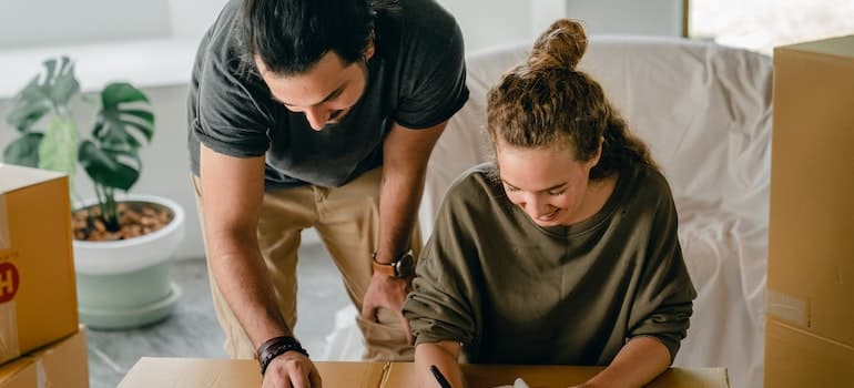 Cheerful diverse couple writing in notebook near boxes before relocation while discussing about the cost of an off-season relocation in Aventura .