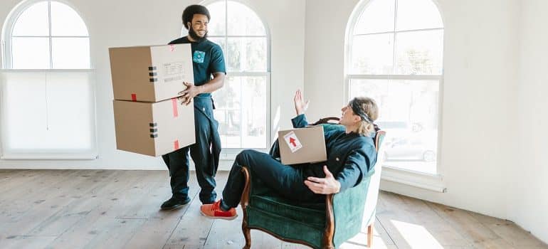 white glove movers, one standing and holding boxes, the other one sitting with a box in his lap