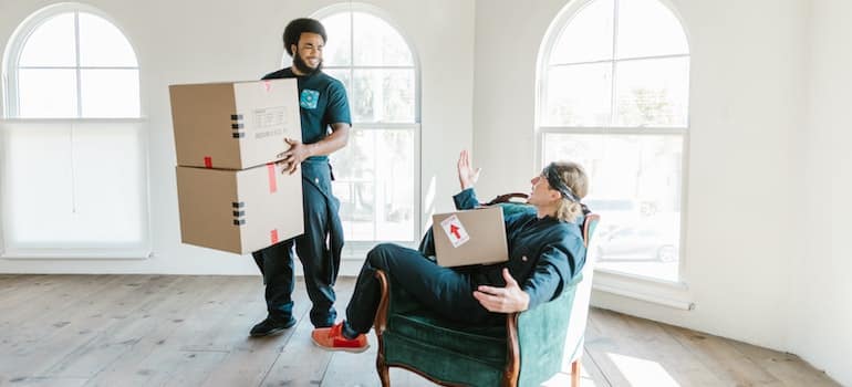 two men standing in a room holding cardboard boxes and talking about who is leaving Miami and why