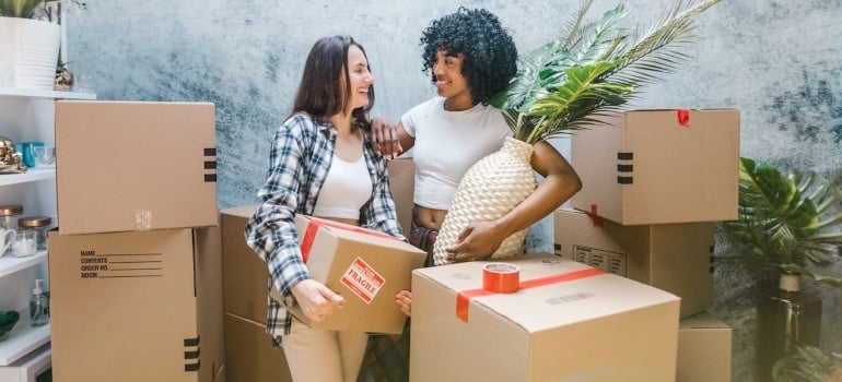 two women, one carrying a box, the other one carrying a plant