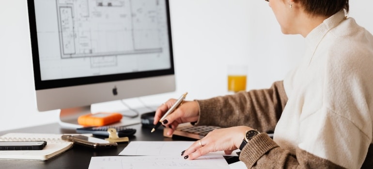 A woman using a computer, paper, and pen planning to pack according to a floor plan.