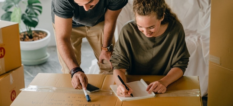 A man assists a woman in packing boxes, while she writes notes on a notepad. Moving boxes are stacked in the background., some being water sport equipment.