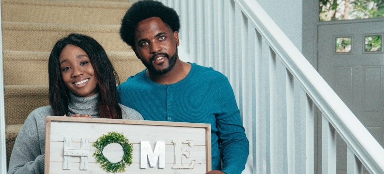 Smiling couple proudly holding a wooden HOME sign after moving into their new place.